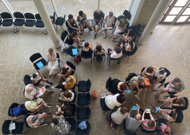 Três grupos de adultos sentados em cadeiras organizadas em círculos, vistos de cima, vários segurando cadernos nas mãos.