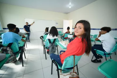 Em uma sala de aula, uma jovem vestindo blusa laranja olha para a câmera, e ao fundo é possível ver um professor dando aula em frente ao quadro.